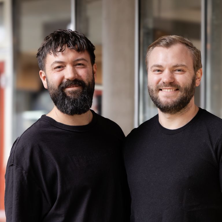 Leo und Anschar in schwarzen T-Shirts stehen nebeneinander und lächeln.