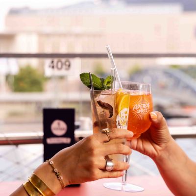 Zwei Hände stoßen mit Cocktails auf der Dachterrasse mit Stadtblick im Hintergrund an.